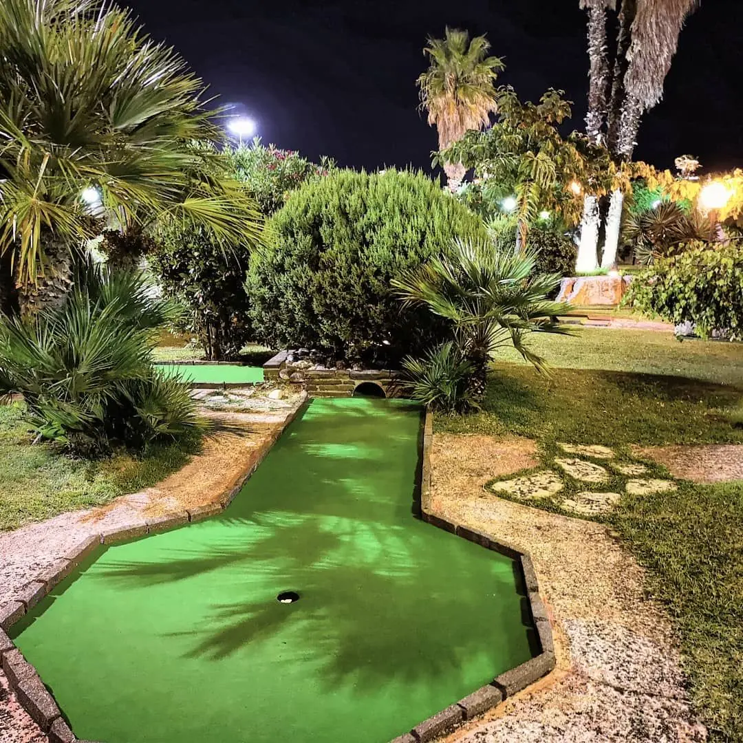 Una pista del minigolf Rivergreen Golf di Rimini illuminata di sera, fra palme e cespugli mediterranei