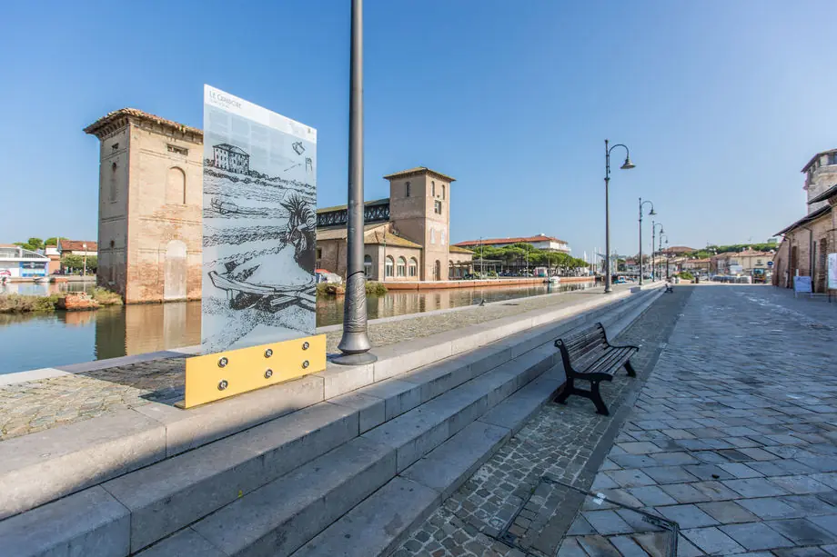 The Cervia canal harbour quay near MUSA, with an illustrated panel, a bench and historic buildings on the opposite bank