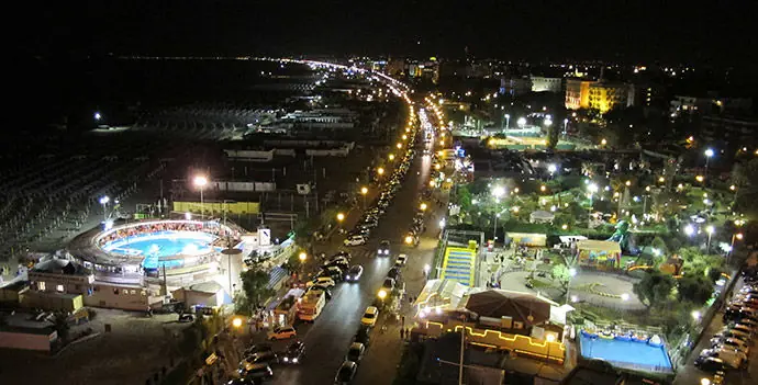 Il lungomare di Rimini di notte visto dall'alto, con il viale illuminato, piscine e giardini a destra e la spiaggia buia