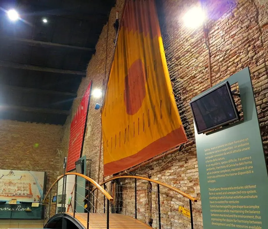 A yellow and red sail hung on the MUSA brick wall, above a wooden walkway, beside a panel with a screen