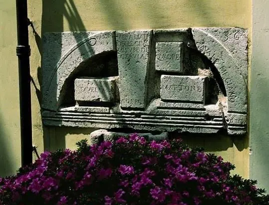 The Pietra delle misure on the yellow wall of Piazza Pisacane, with the tavella, quadrè and brick panels above purple flowers