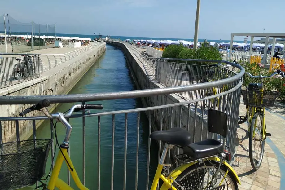 Bicycles leaning against the railings of a channel descending towards Misano Adriatico beach, with parasols in the distance