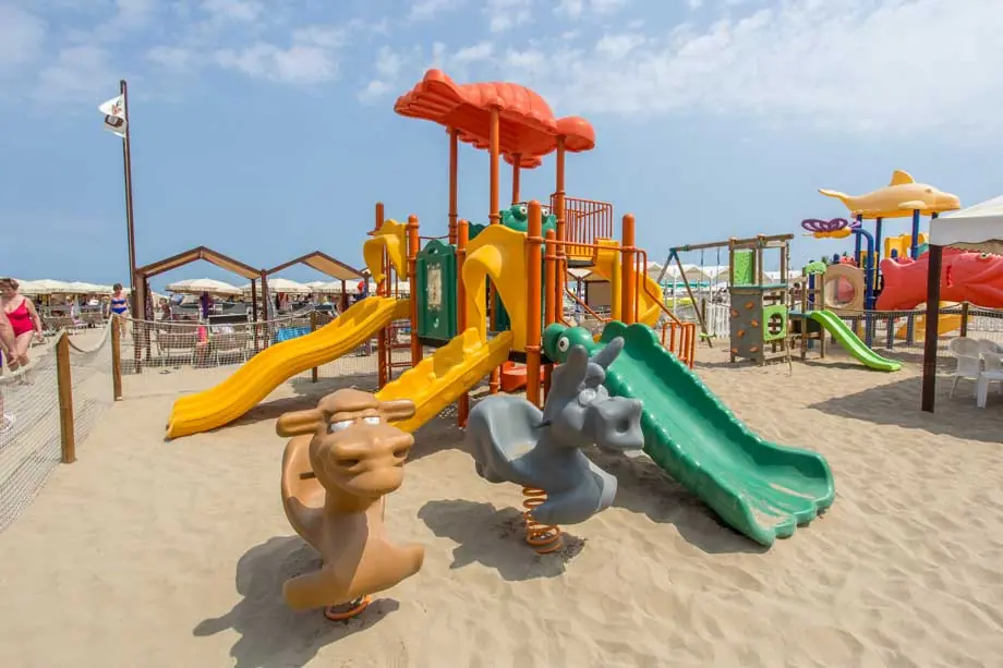 Gelbe und grüne Rutschen, eine Kamelwippe und weitere bunte Spielgeräte im Sand eines Strands von Cattolica