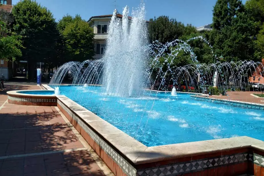 Ein Brunnen mit hohen Wasserstrahlen und blauem Becken auf einem Platz in Cattolica, zwischen Bäumen und Häusern