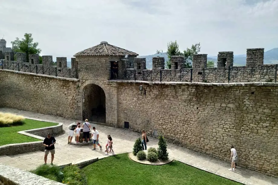 Le mura merlate in pietra di una fortezza di San Marino, con un cortile erboso e i visitatori sul selciato