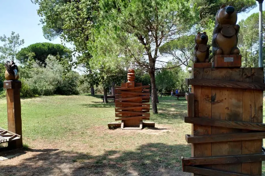 Sculptures en bois en forme d'animaux entre les pins du Parco Naturale di Cervia, avec des personnes sur les bancs au fond de la pelouse