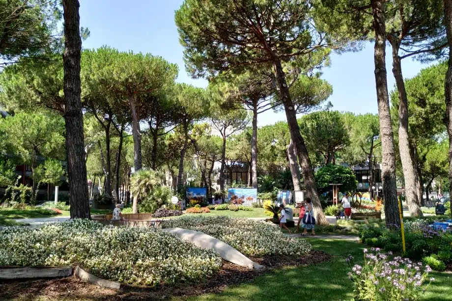 Ein blühendes Beet im Schatten der Pinien auf einem der Kreisverkehre von Milano Marittima, mit einigen Passanten im Hintergrund