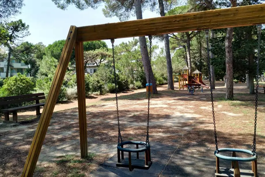 Schaukeln für kleine Kinder in einem Park im Schatten der Pinien in Milano Marittima, mit Bank und weiteren Spielgeräten im Hintergrund