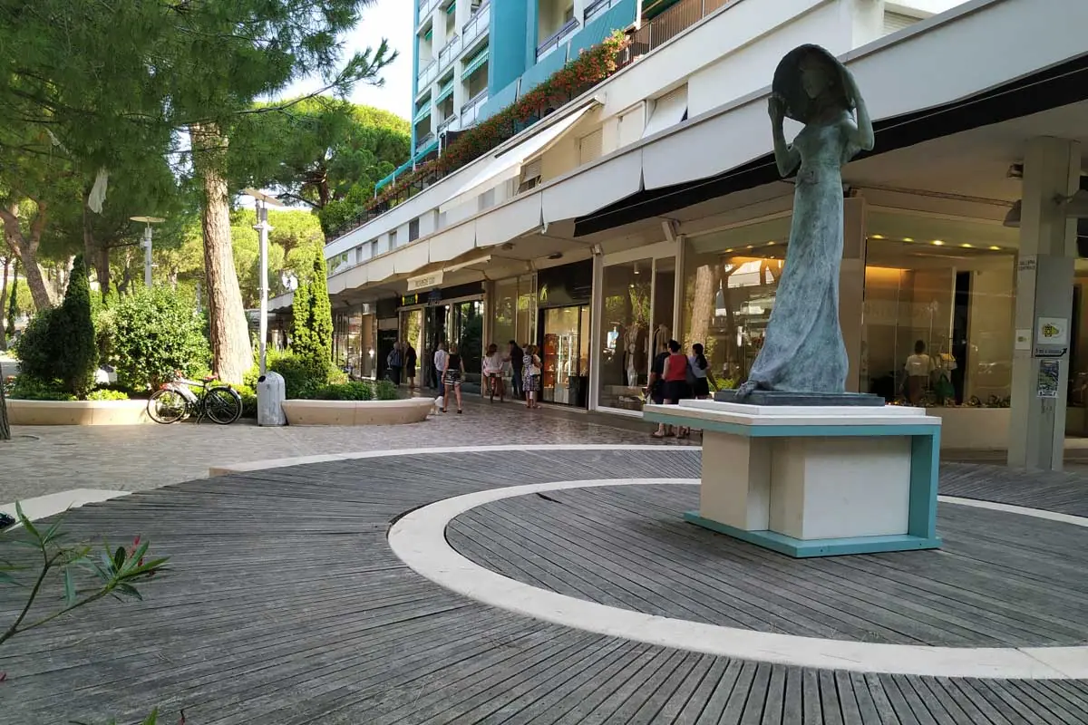 Eine Fußgängerzone im Zentrum von Milano Marittima mit Schaufenstern unter Arkaden, Pinien und einer Bronzestatue in der Mitte