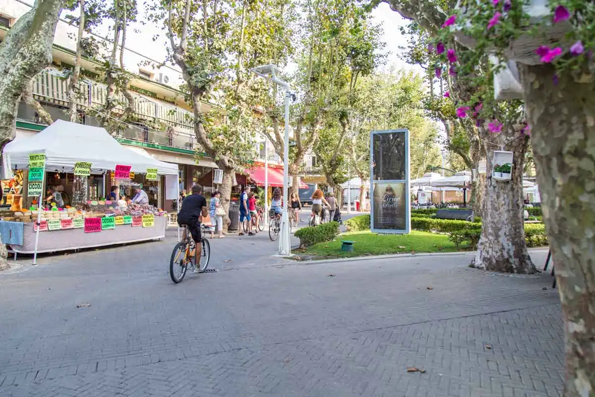 Ein Stand mit regionalen Erzeugnissen unter den Platanen der Fußgängerzone von Bellaria Igea Marina, mit Passanten und Fahrrädern