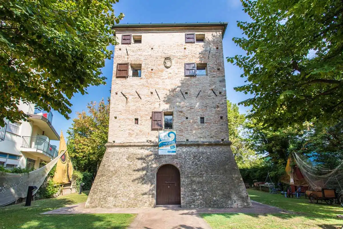 La Torre Saracena di Bellaria, una torre in mattoni con la base a scarpa e il portone ad arco, fra gli alberi del giardino