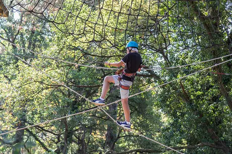 A boy wearing a helmet and harness walks along a rope between the trees, holding the ropes at the sides