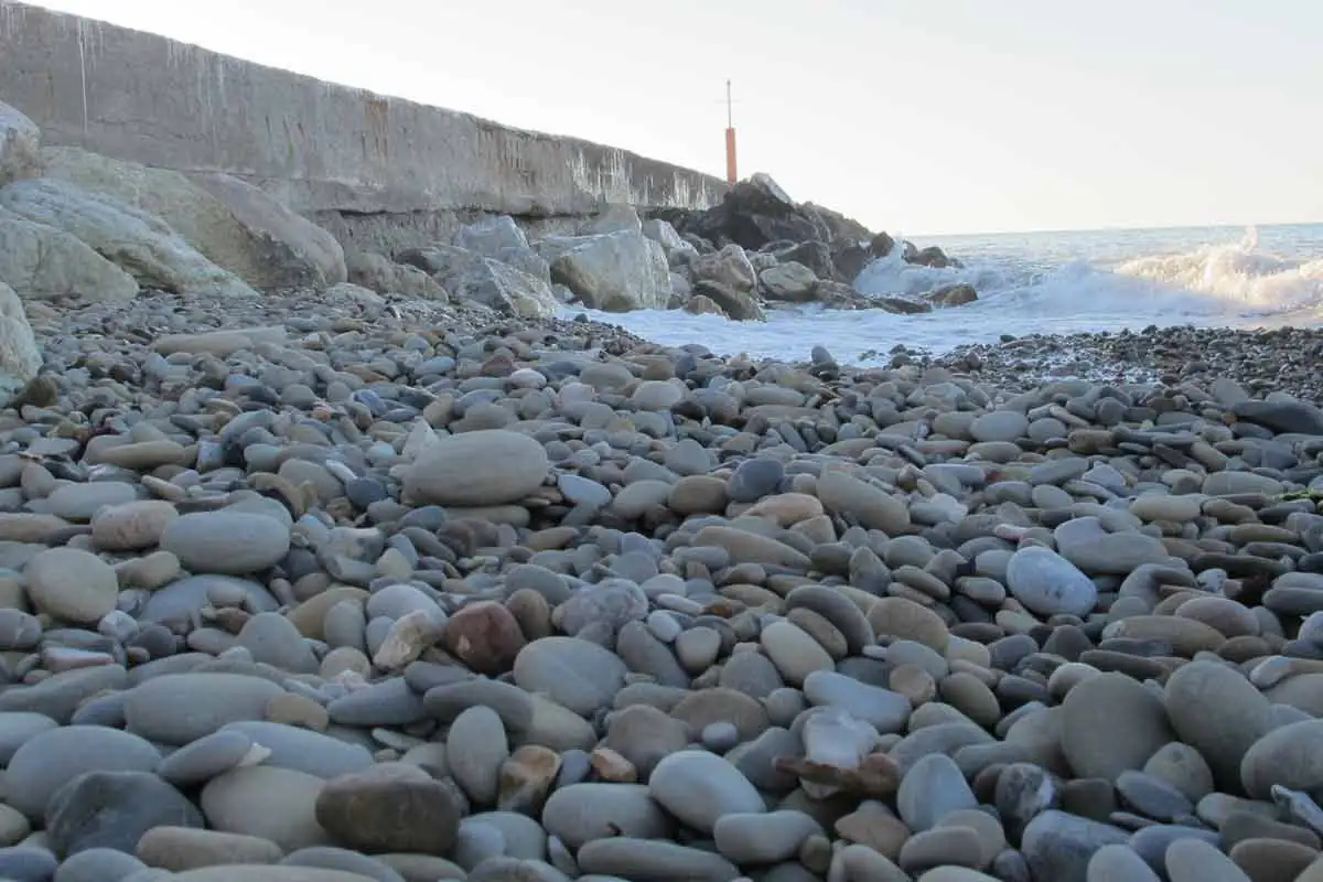 Vom Meer geglättete Kiesel an einem Strand von Gabicce Mare unter einer Kaimauer mit Wellen im Hintergrund