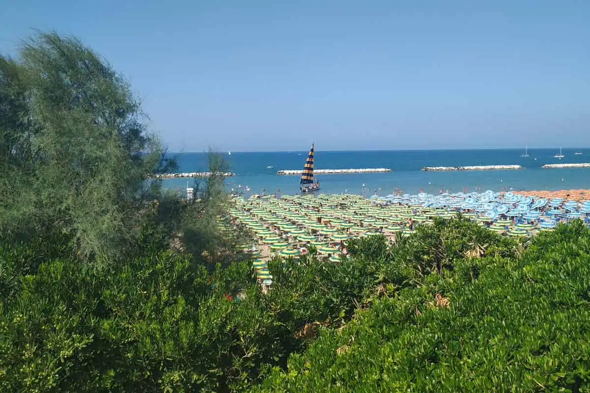 Der Strand von Gabicce Mare von oben zwischen Vegetation mit Sonnenschirmen, vorgelagerten Wellenbrechern und einem Segelboot