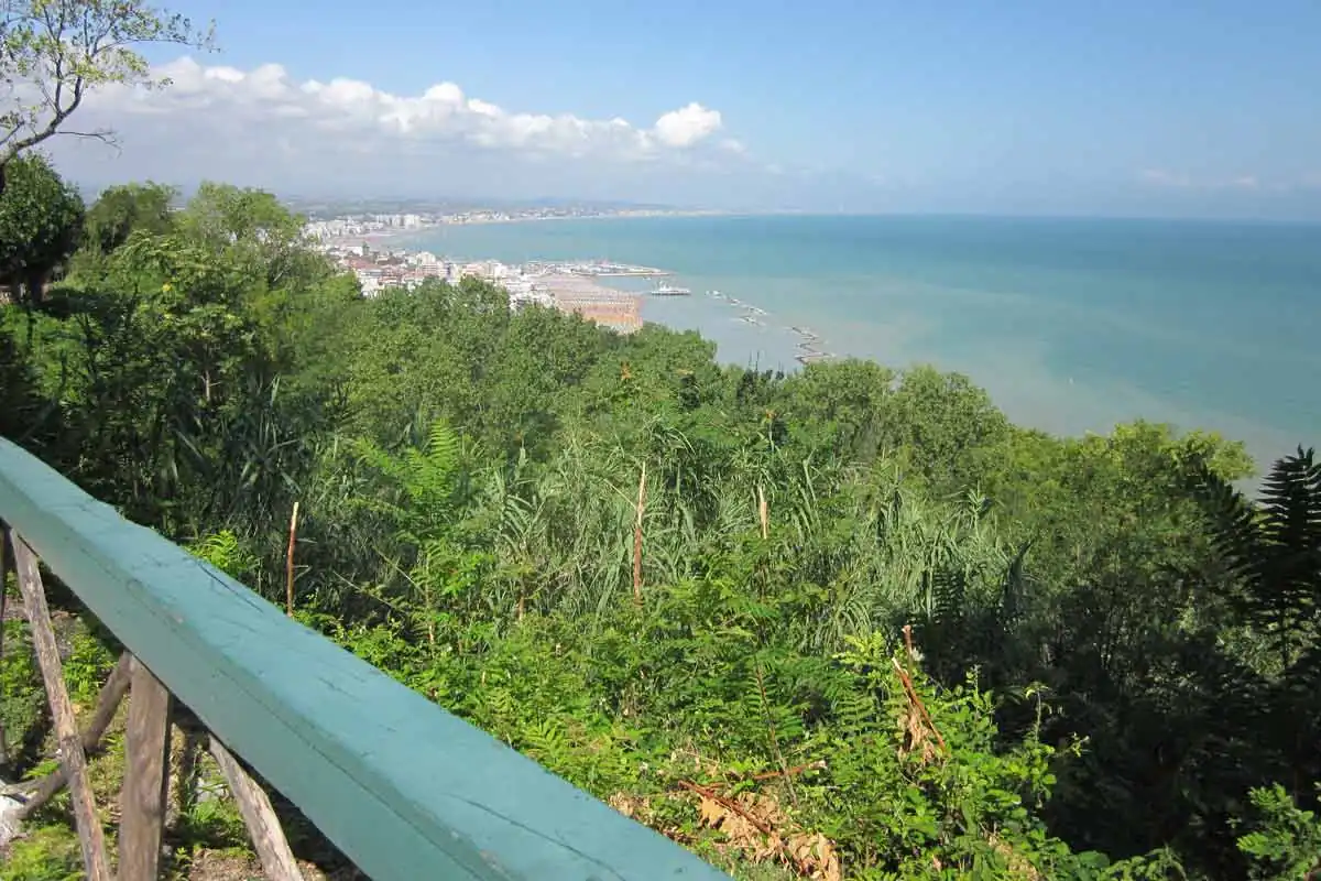 La costa vista da Gabicce Monte oltre una staccionata verde, con il promontorio coperto di macchia e la baia fino a Cattolica