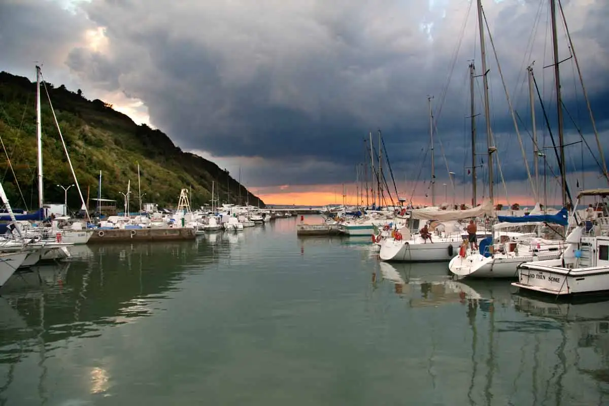 Barche a vela ormeggiate nel porticciolo di Vallugola, sotto il promontorio boscoso e un cielo di temporale
