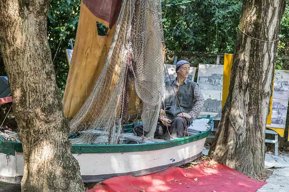 A fisherman mannequin seated in a boat with nets and a sail in the Torre Saracena garden, in front of old photographs
