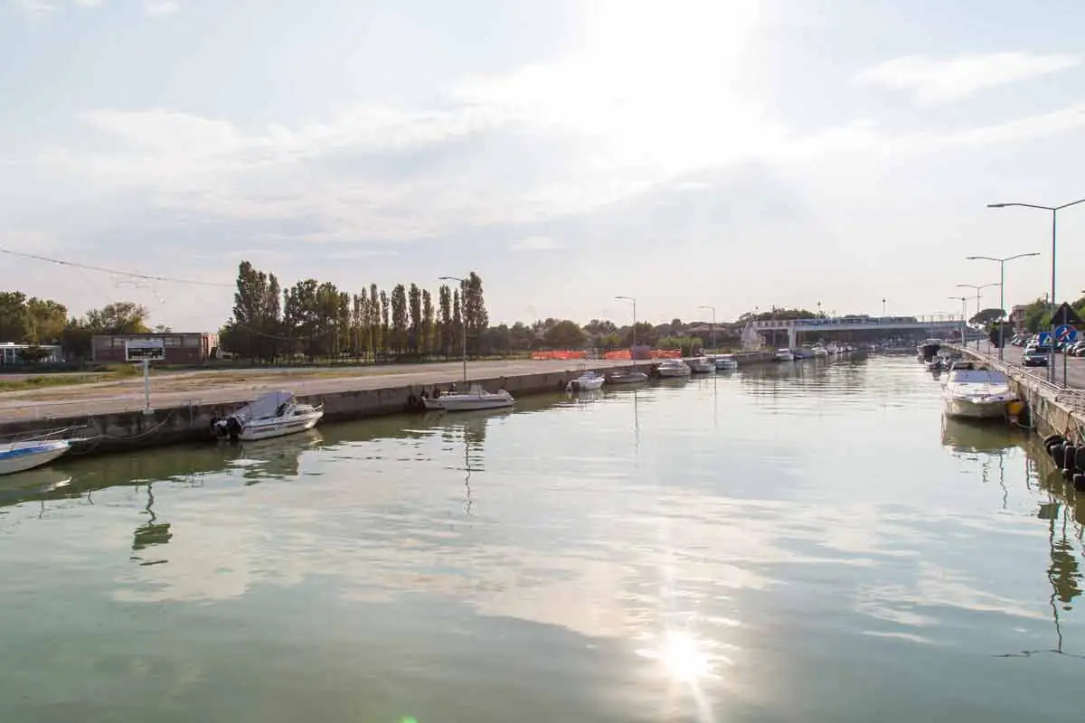 Bellaria Igea Marina's canal harbour with a few moored boats and a row of pines at the far end, backlit by the sun