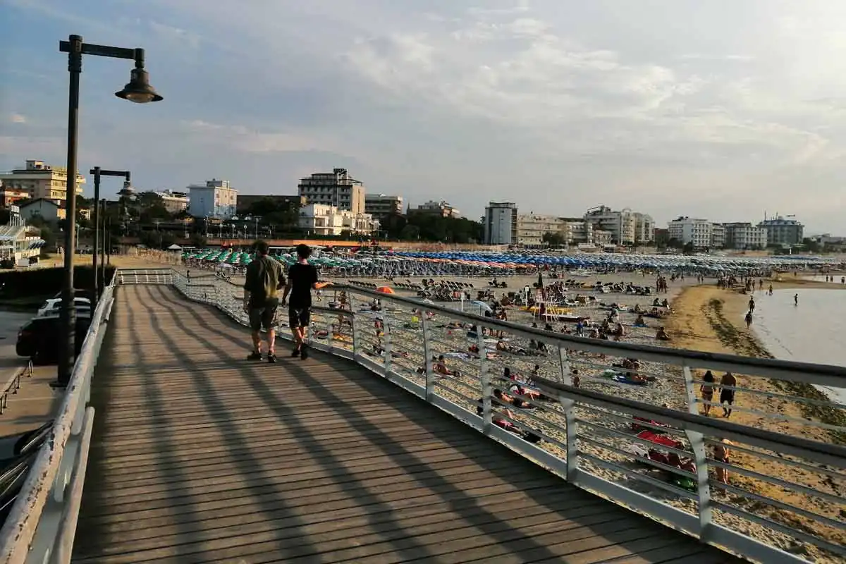 Une passerelle en bois sur la plage de Cattolica au couchant, avec baigneurs sur le sable et parasols en arrière-plan