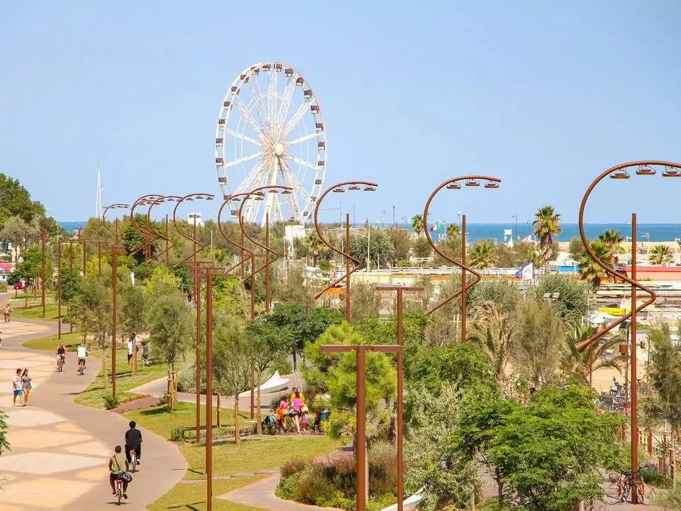 I viali del Parco del Mare a Rimini, con ciclisti e pedoni fra gli alberi, lampioni ad arco e la ruota panoramica sul fondo