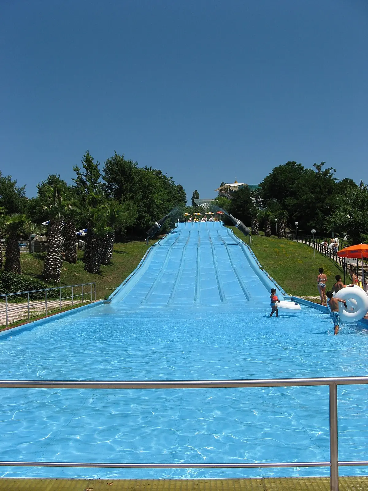 Un largo scivolo azzurro a più corsie che scende in una vasca, tra palme e prati, nel parco acquatico di Riccione