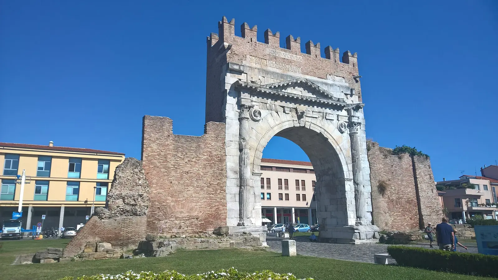 L'Arco di Augusto di Rimini visto di tre quarti, con il coronamento merlato, le mura in laterizio ai lati e un cielo azzurro