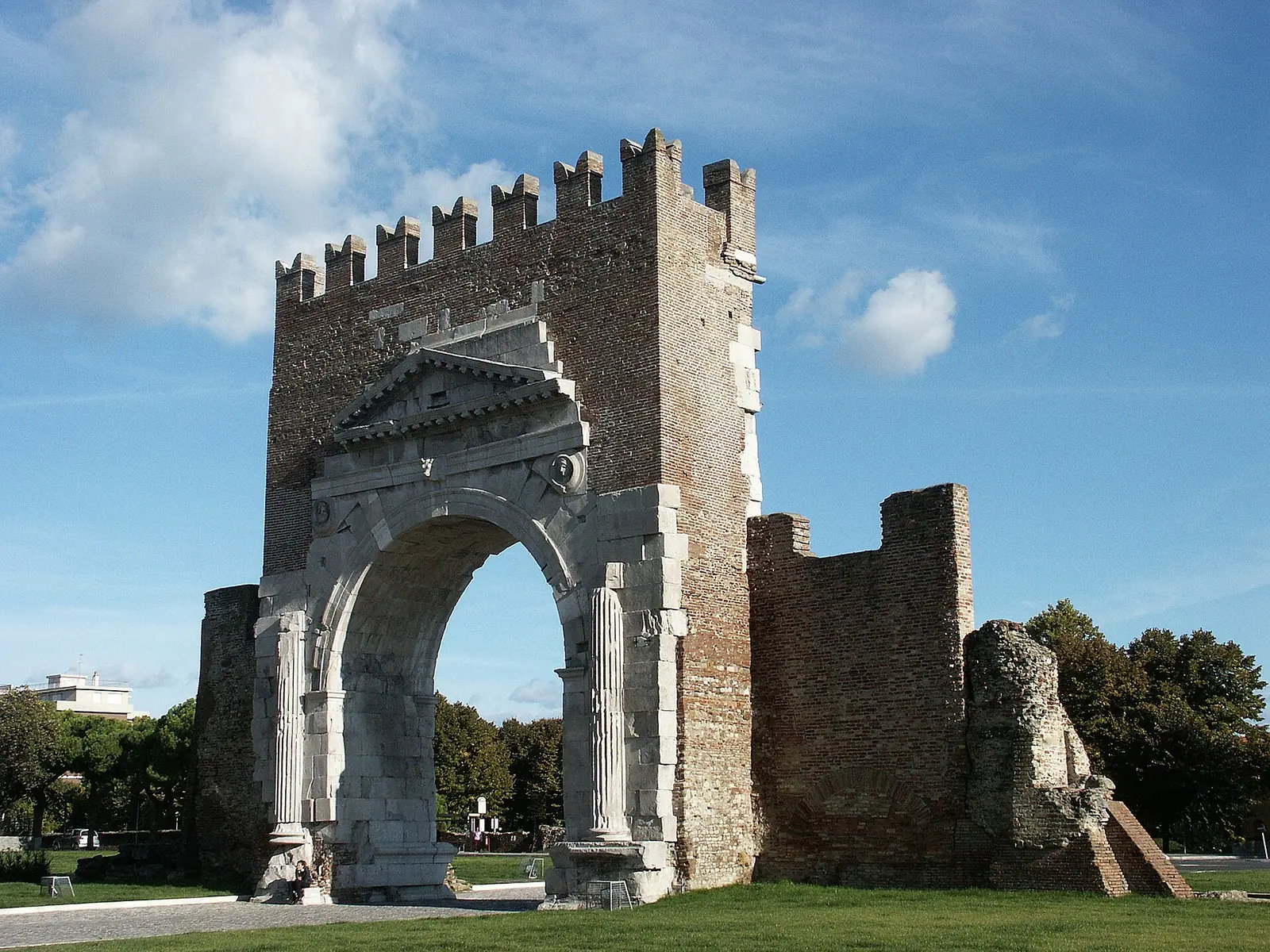 Arco di Augusto in Rimini seen from the side across the lawn, with brick battlements and the adjoining wall section