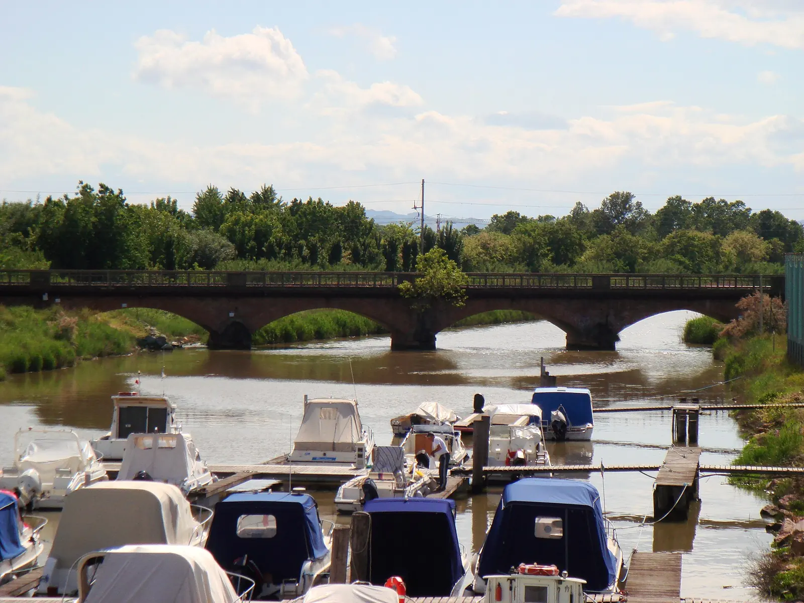 An arched brick bridge over the canal, with boats moored at jetties in the foreground and greenery along the banks