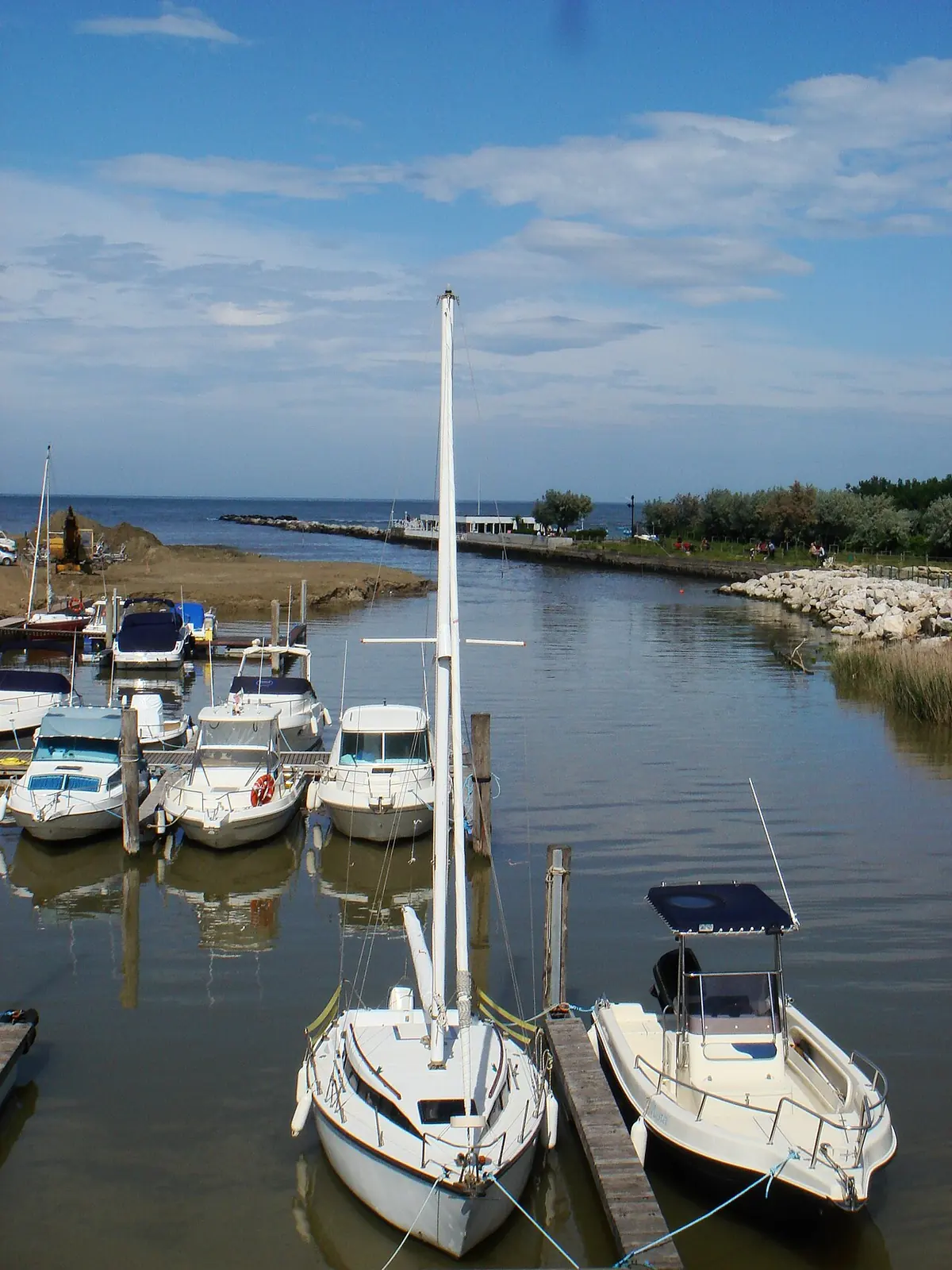 Barche a motore e a vela ormeggiate ai pontili di un canale, con il molo e il mare sul fondo