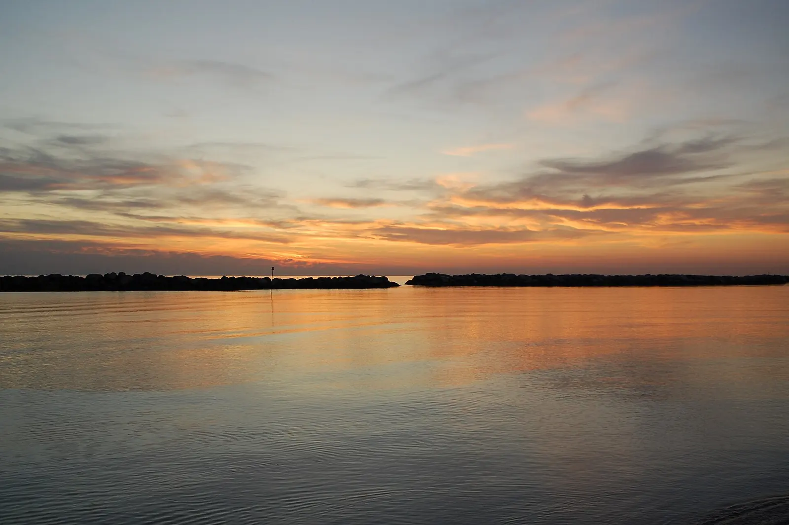 Ruhiges Meer unter orange-blauem Himmel, mit zwei Wellenbrechern als Silhouetten