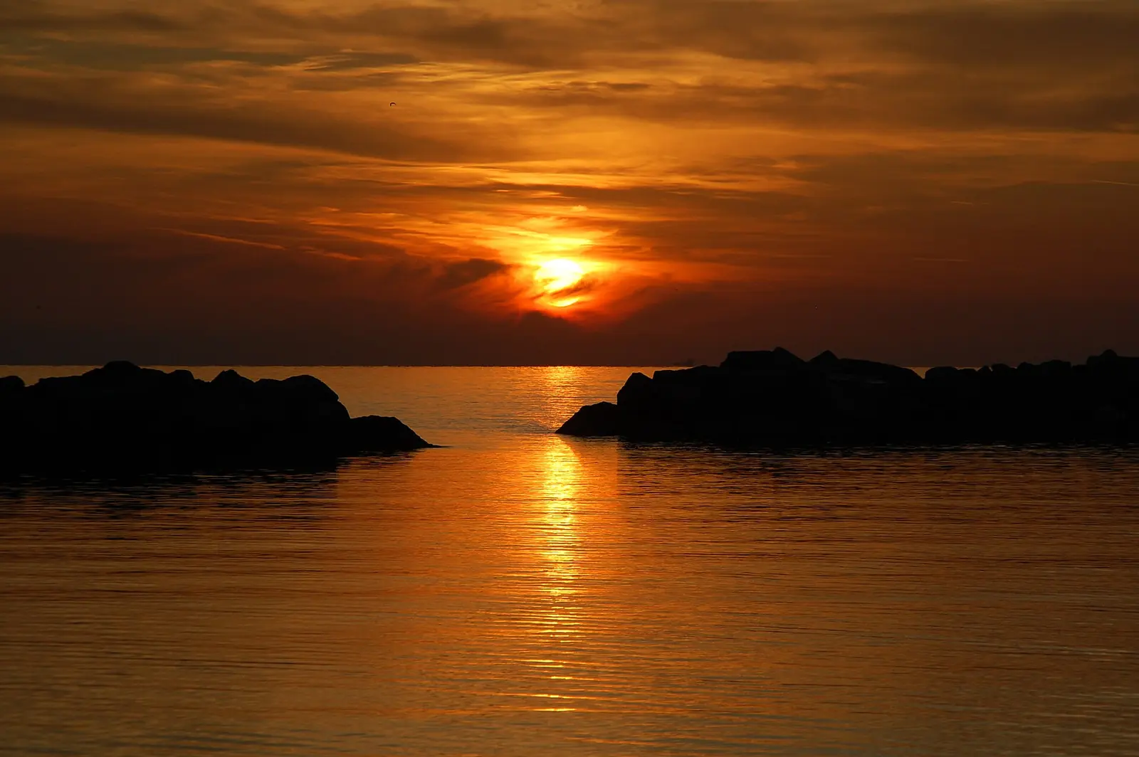 The low sun on the horizon and its reflection on calm sea, between two silhouetted breakwaters