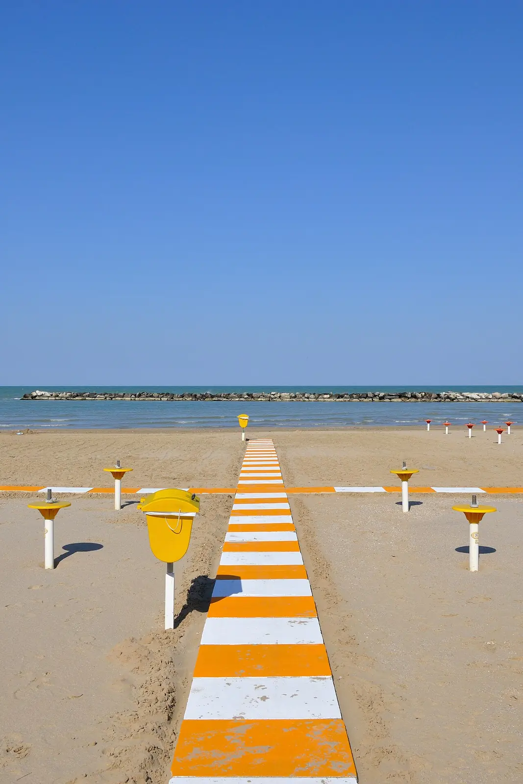 Ein orange-weiß gestreifter Steg am Strand mit Sonnenschirmstangen und einem Wellenbrecher am Horizont