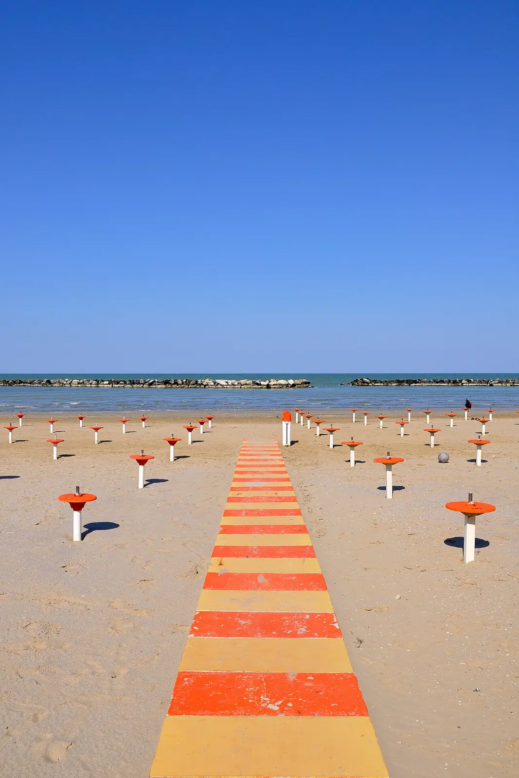 An orange and yellow striped walkway crosses the sand towards the sea, with a breakwater before the horizon