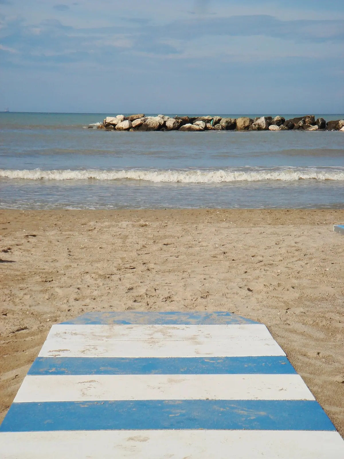 A white and blue striped walkway on the sand and a breakwater in front of the calm sea