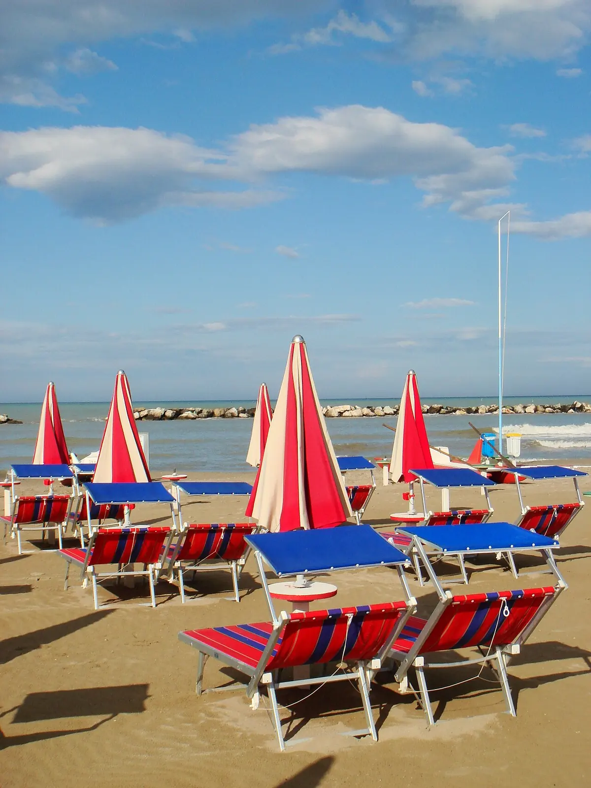 Red and blue striped loungers and closed red and white parasols on the sand, with a breakwater facing the sea