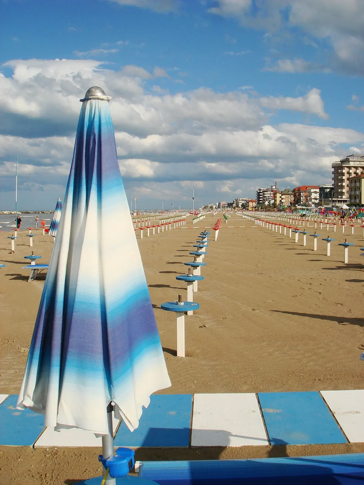 A closed parasol in shades of blue in the foreground, with the long beach and aligned poles stretching to buildings at the far end