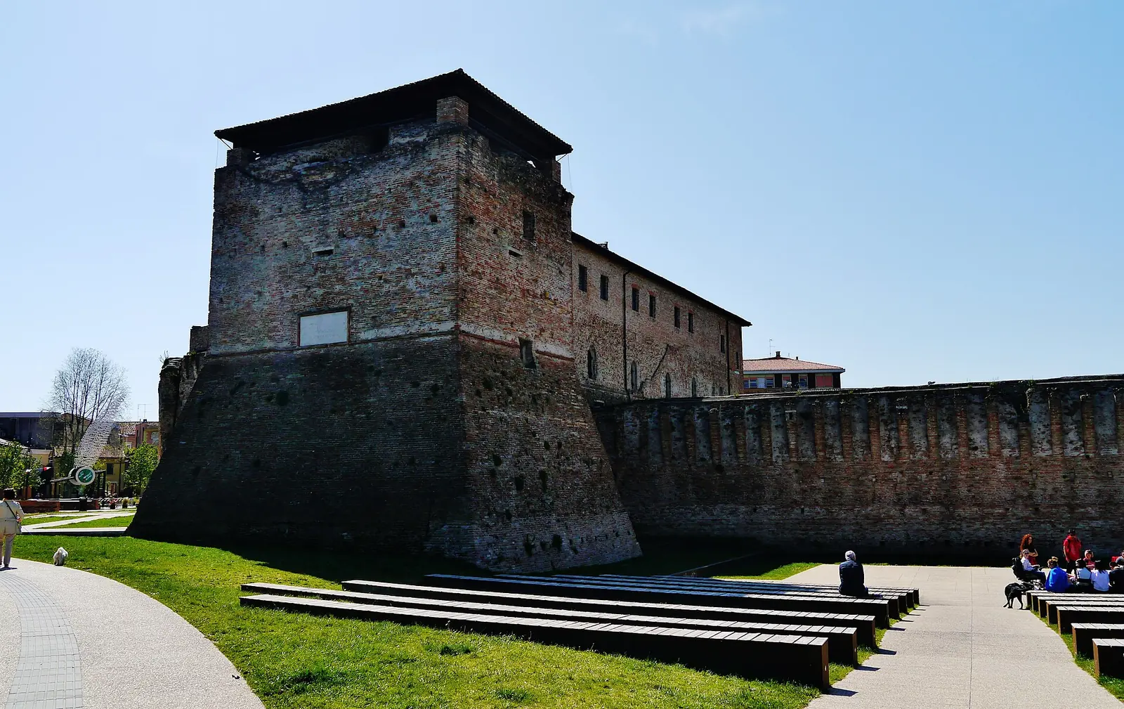 Die Westseite des Castel Sismondo in Rimini mit Backsteinturm, Mauern und Holzstufen auf dem Rasen davor