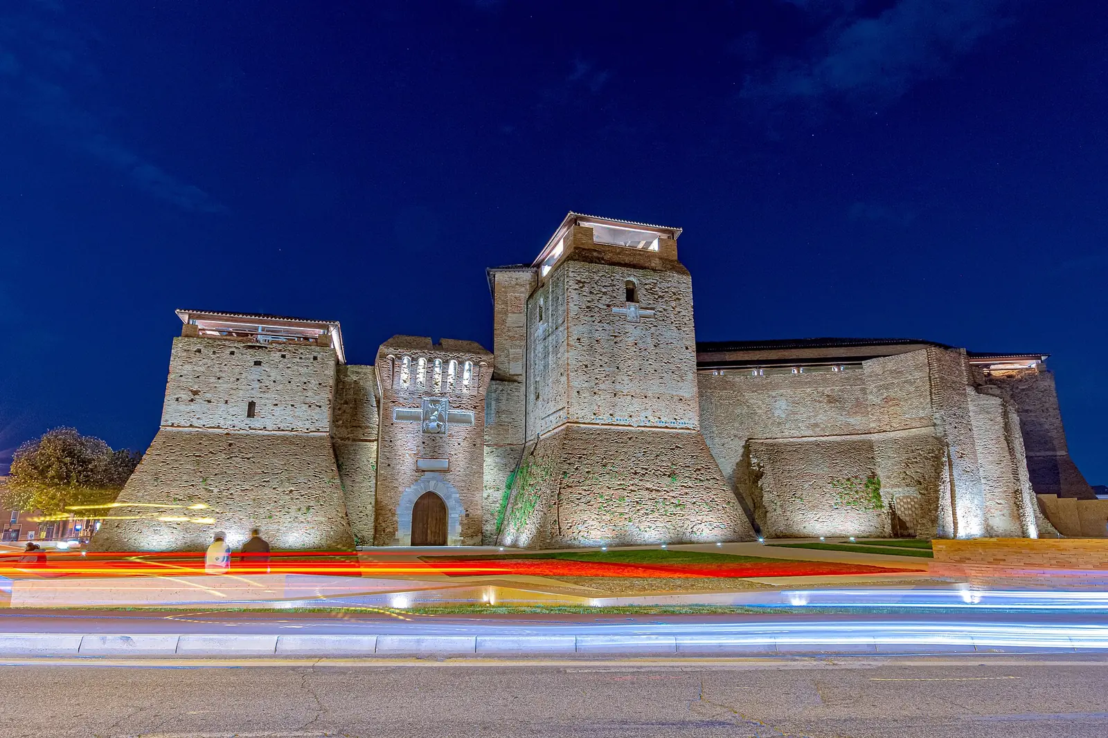 Castel Sismondo a Rimini illuminato di sera, con le torri a scarpa e le scie luminose delle auto in primo piano