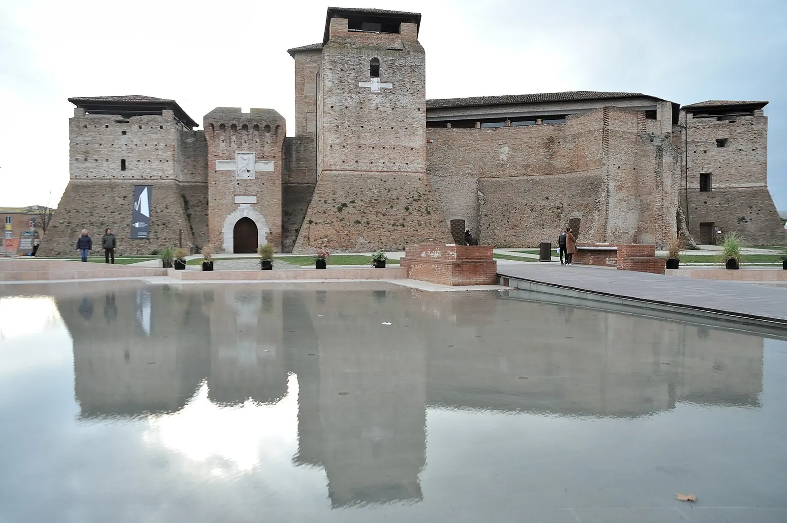 Castel Sismondo a Rimini riflesso nel velo d'acqua di piazza Malatesta, con il rivellino, il mastio e alcuni passanti lontani