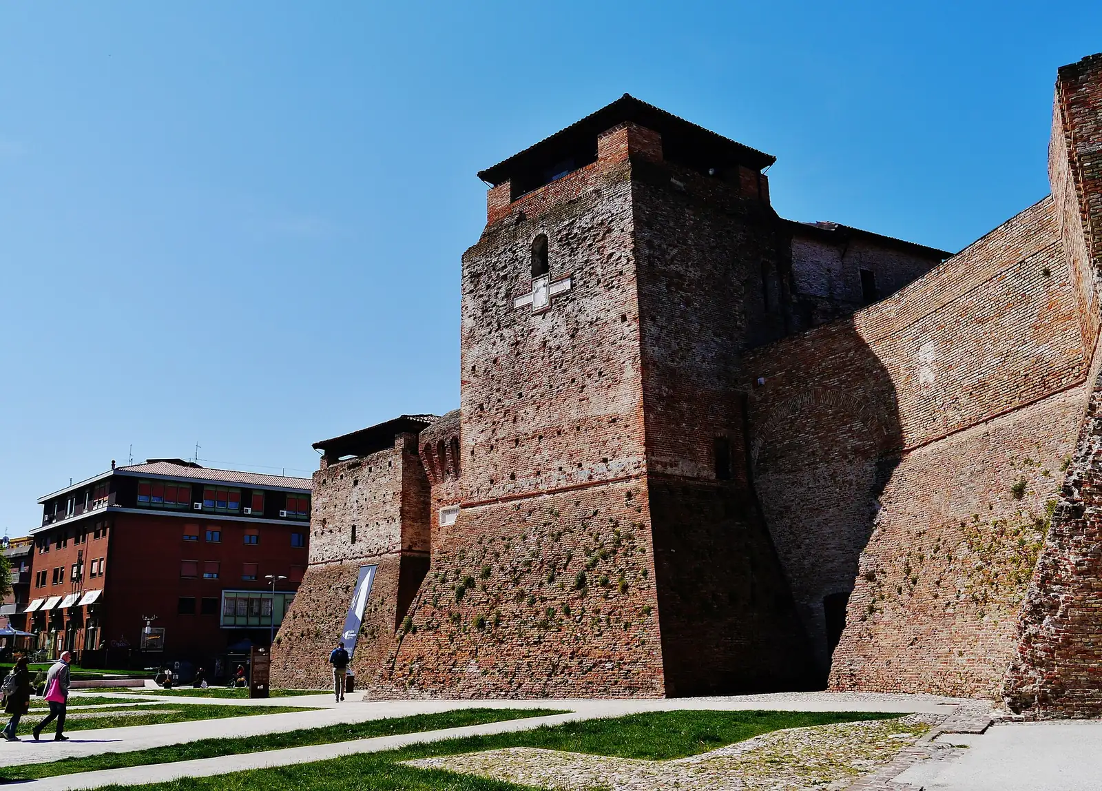 Der zinnenbekrönte Turm und die geböschten Mauern des Castel Sismondo in Rimini unter klarem Himmel, links ein rotes Gebäude