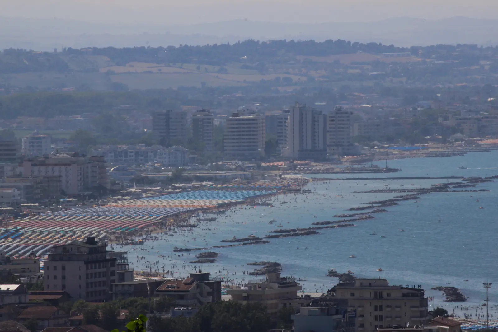 Cattolicas Küste von oben mit Sonnenschirmreihen, parallelen Wellenbrechern und Gebäuden hinter dem Strand