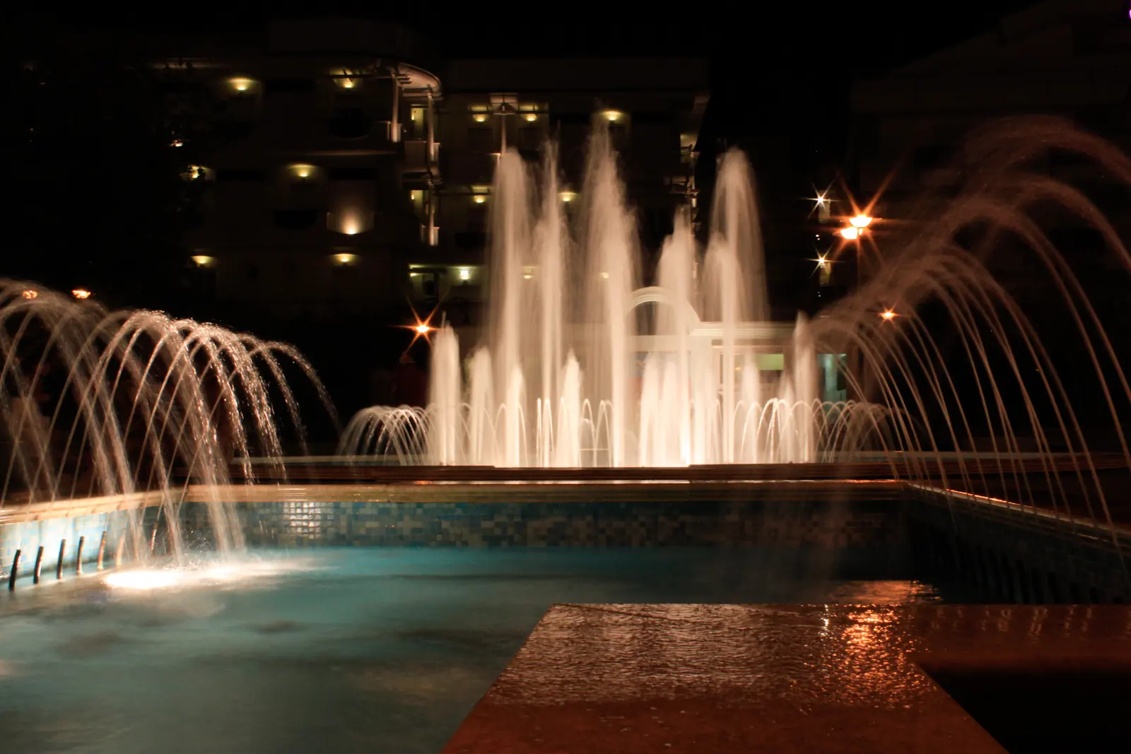 Getti d'acqua illuminati di una fontana di Cattolica di notte, con un edificio dalle finestre accese sullo sfondo