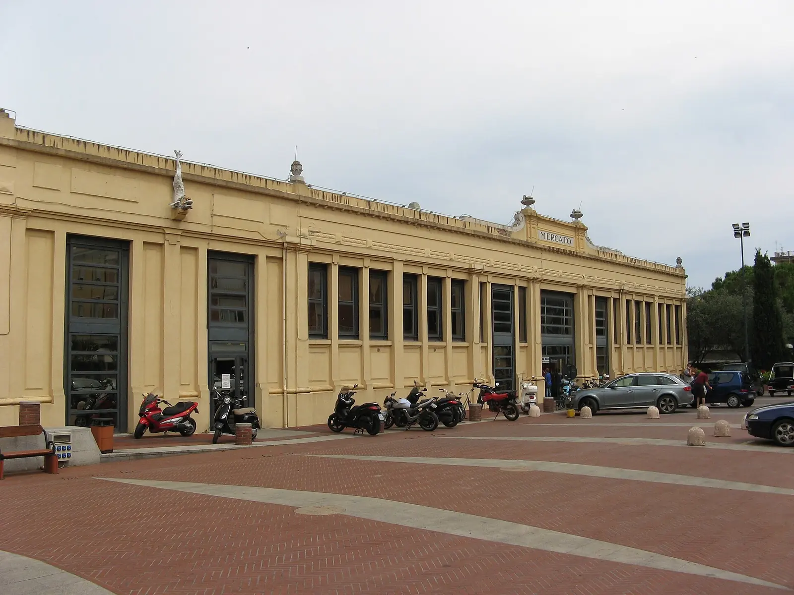 La facciata gialla del Mercato Coperto di Cattolica, con grandi vetrate, motorini parcheggiati e il piazzale in mattoni