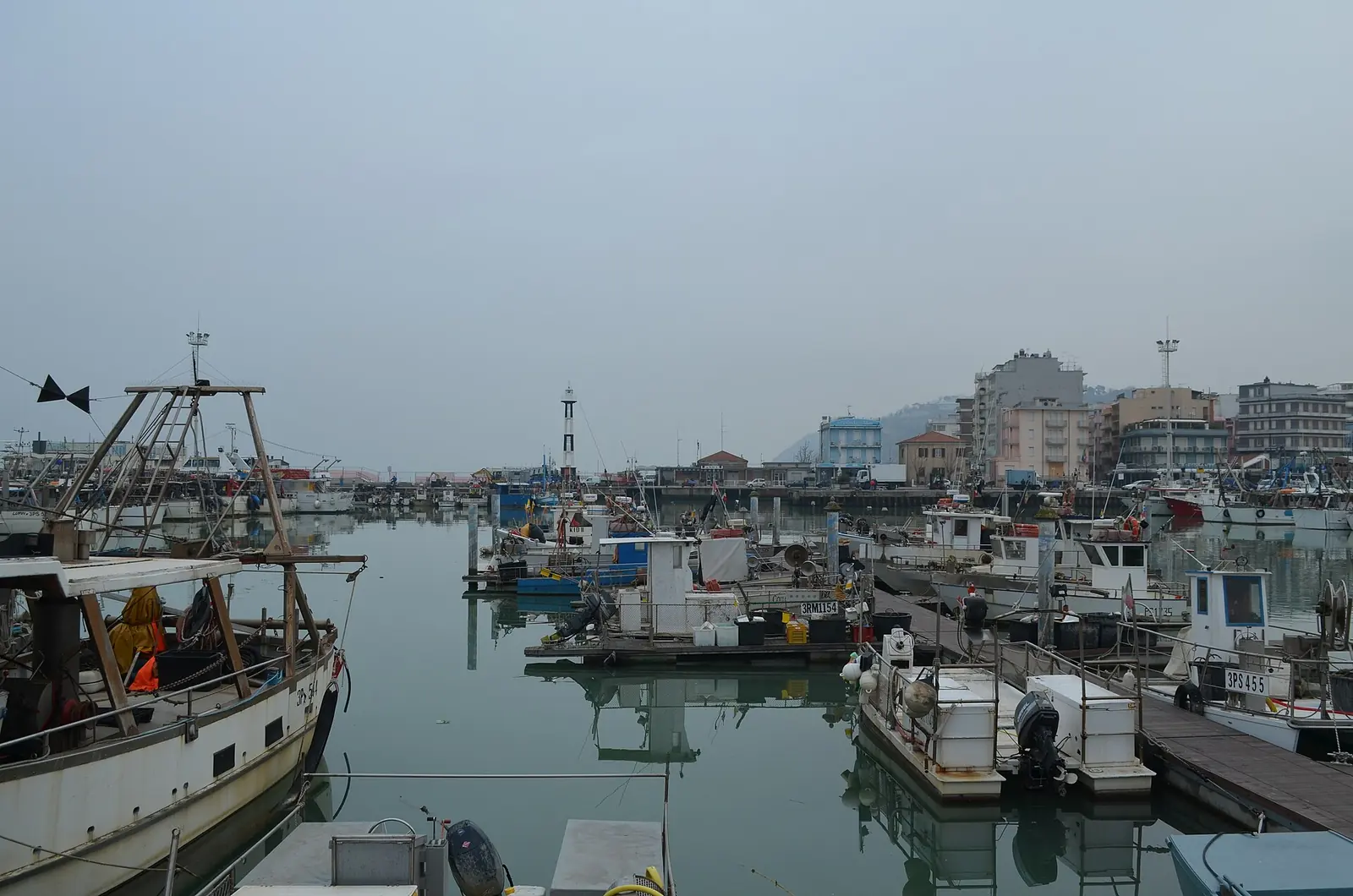 Pescherecci ormeggiati nel porto canale di Cattolica, con il faro bianco e nero e le case sul fondo