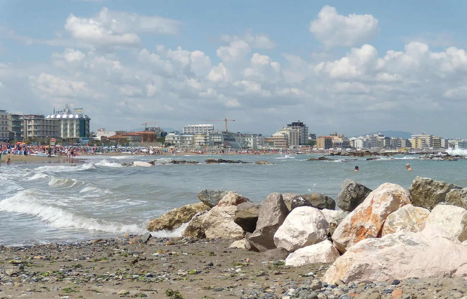 Onde che si frangono fra i massi di una scogliera, con la spiaggia affollata e gli edifici di Cattolica sullo sfondo
