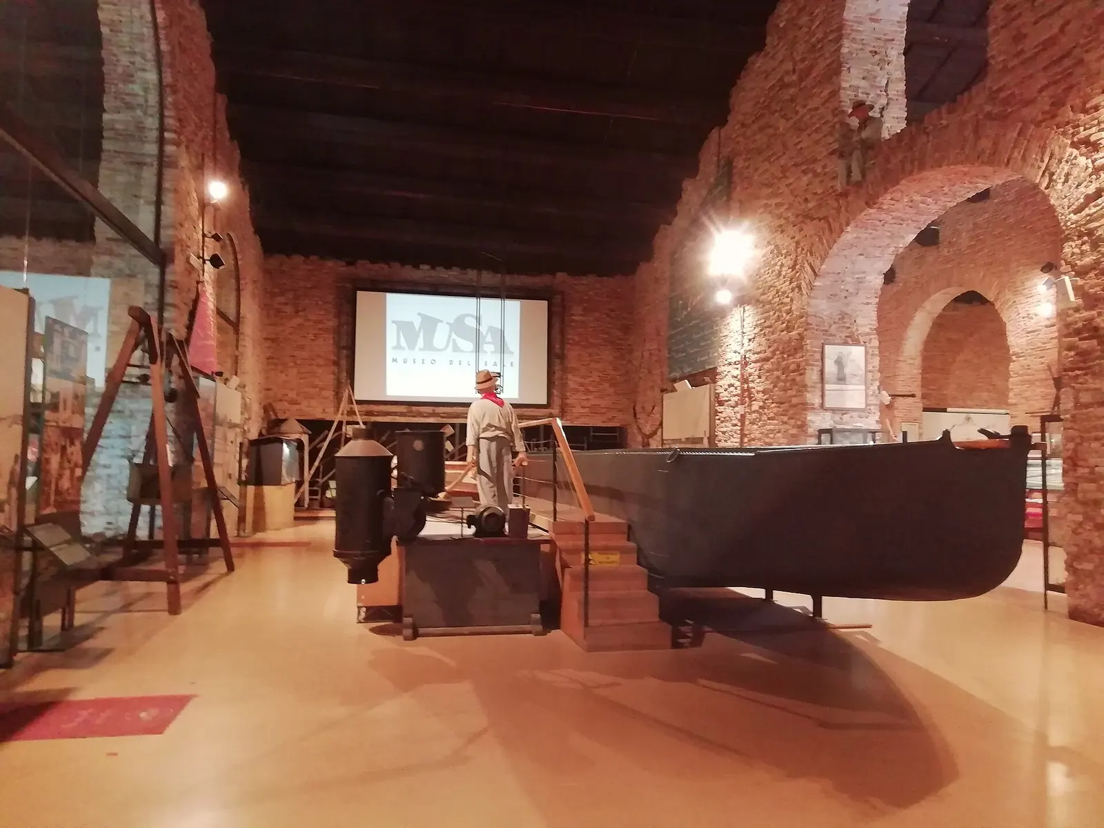 A room at MUSA in Cervia with brick vaults, a boat on display and a screen showing the museum logo