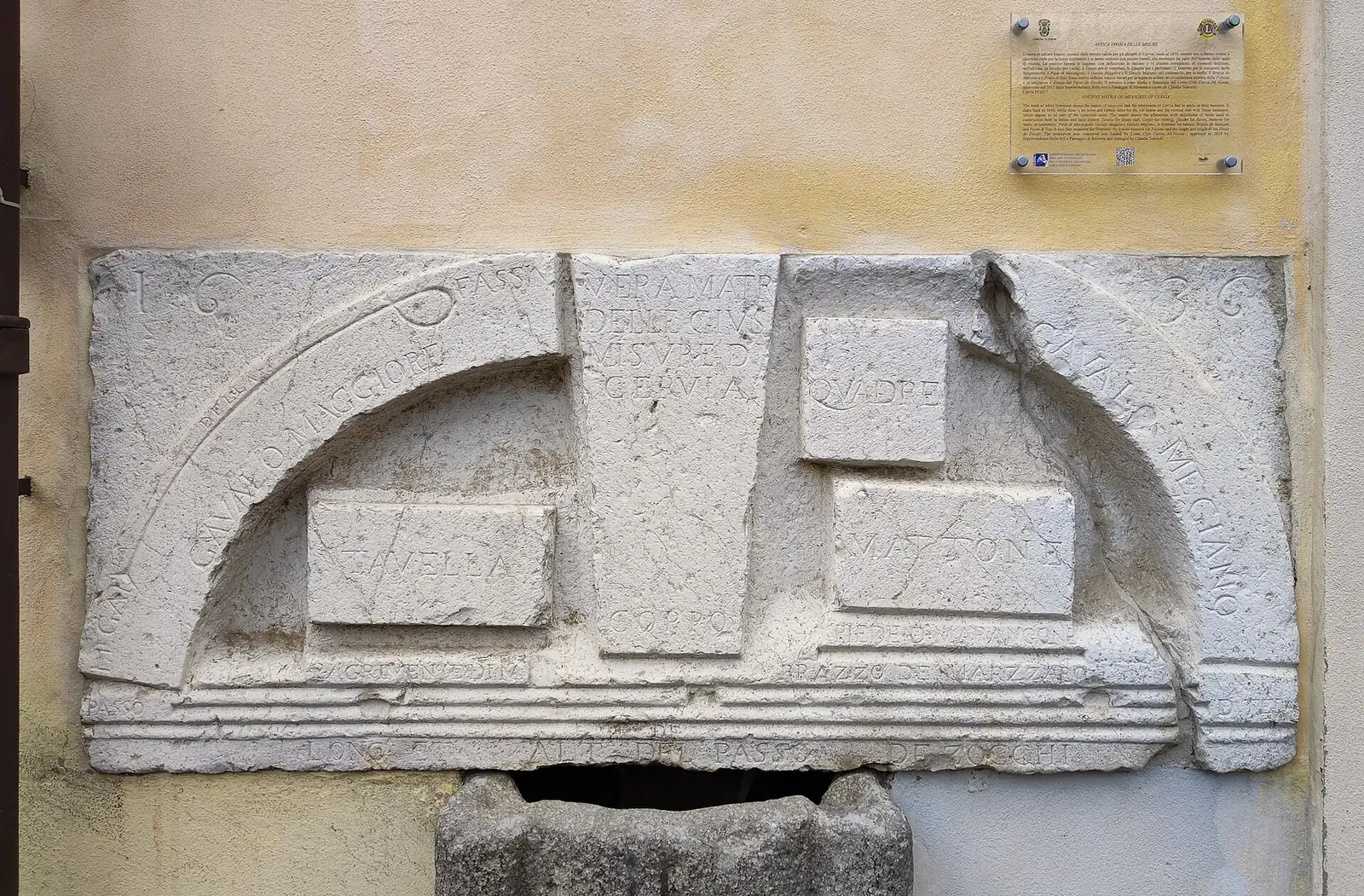 The measuring stone in Cervia, a white limestone slab with engraved units of measurement, set into a yellow wall