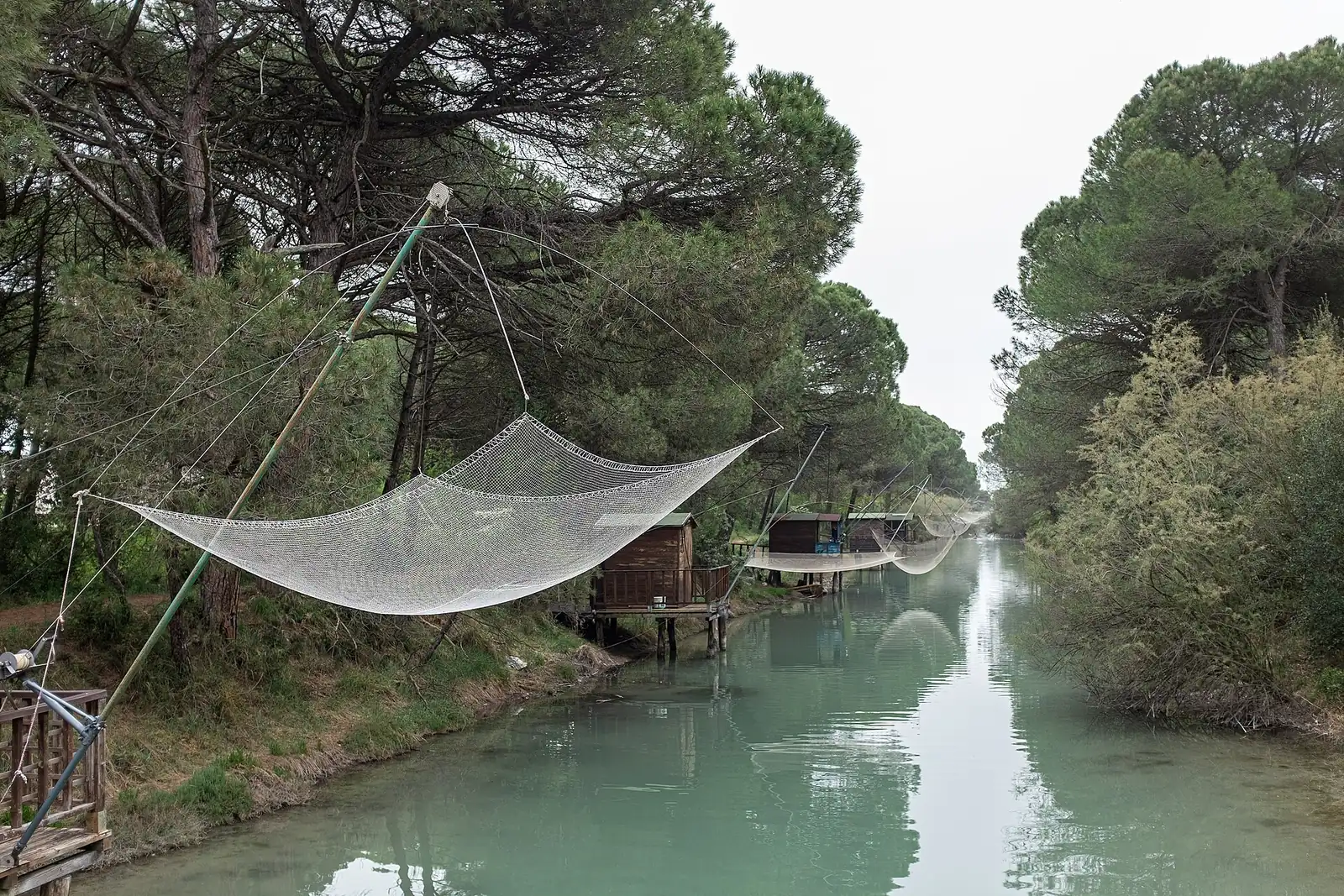 Le Canalino entre les pins, avec un grand filet de pêche relevé et des cabanes sur pilotis sur la rive