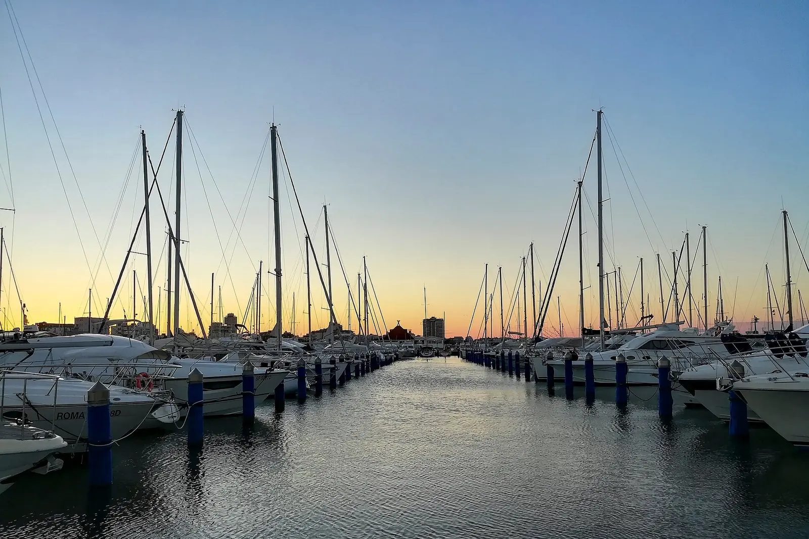 Vertäute Boote zwischen blauen Pfählen in einem Yachthafen von Cervia unter orangefarbenem Abendhimmel