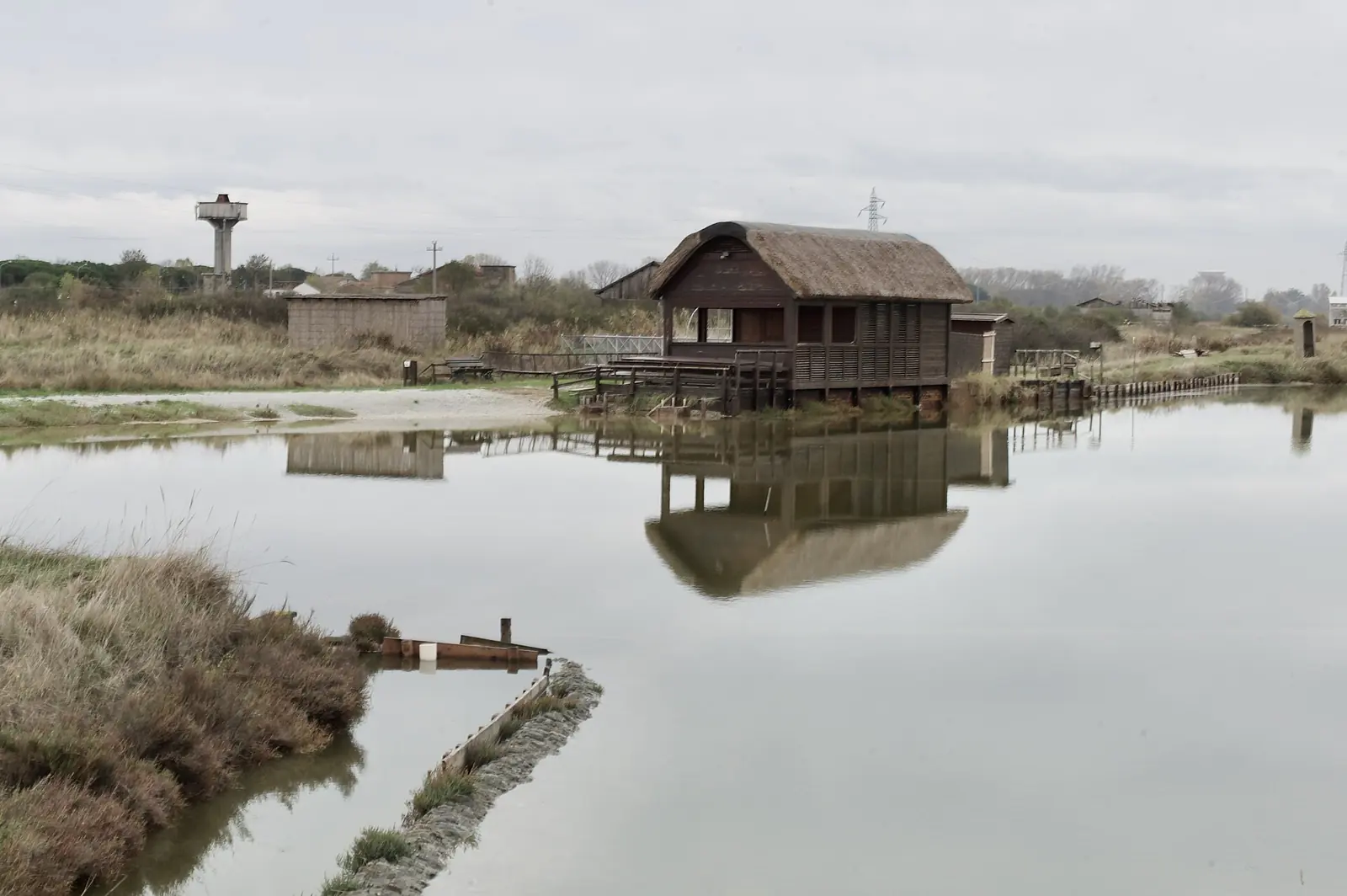 A wooden hut with a reed roof over the water at Salina Camillone in Cervia, with a tank in the background